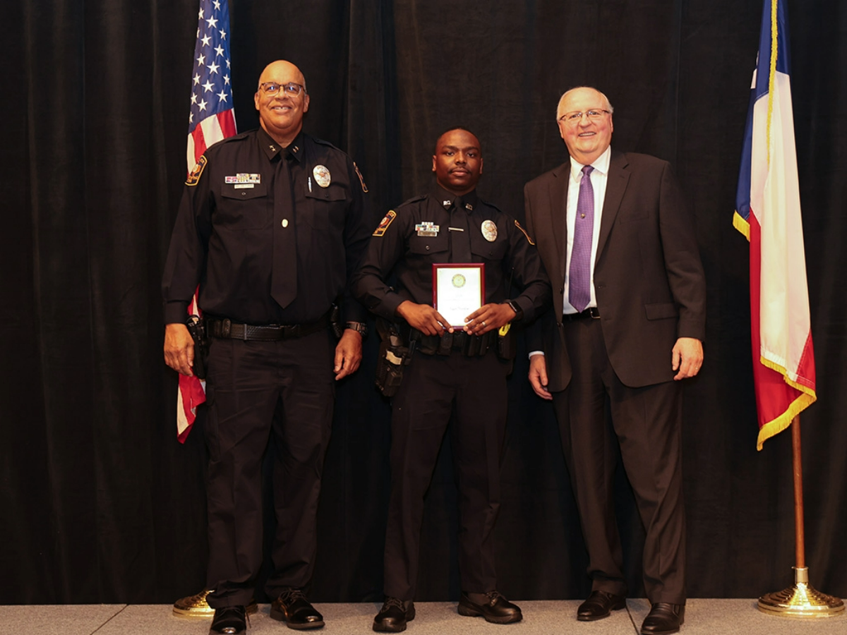 Three men pose onstage at an awards ceremony—an officer in the center holding a plaque—flanked by a uniformed UT Police leader (left) and a suited administrator (right), with U.S. and Texas flags on either side.